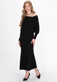 Rochie tricotată neagră cu design off-the-shoulder, mâneci lungi și tiv evazat. Asortată cu tocuri negre cu vârf ascuțit. Textură netedă.