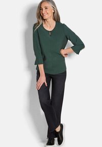 Grünes, langärmliges Oberteil mit einem Schlüsselspalt-Detail, kombiniert mit dunklen Jeans und schwarzen Schuhen. Das Oberteil zeichnet sich durch eine dezente Textur und einen strukturierten Schnitt aus.