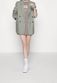 Blazer grigio a quadri con due bottoni neri, indossato sopra una maglietta marrone. Abbinato a sneaker bianche e calze bianche al ginocchio.