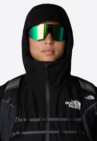 Czarna wodoodporna kurtka z kapturem, srebrne i zielone okulary odblaskowe, szaro-czarne paski na ramię oraz białe akcenty logo.