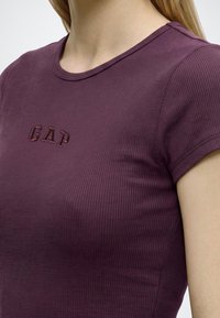 Κοντινό πλάνο ενός ατόμου που φοράει μια ριμπ κοντομάνικη μπλούζα σκούρου μοβ χρώματος με τη λέξη "GAP" κεντημένη στο στήθος.