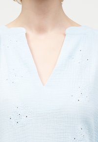 Κοντινό πλάνο λαιμού και άνω μέρους στήθους με ανοιχτό μπλε υφασμάτινο ένδυμα με κεντητά ανθικά σχέδια και λαιμόκοψη σε σχήμα V.