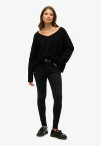 Czarny, dziany, oversize sweter z dekoltem w kształcie V, w połączeniu z czarnymi dopasowanymi dżinsami i czarnymi butami o strukturalnym wykończeniu.