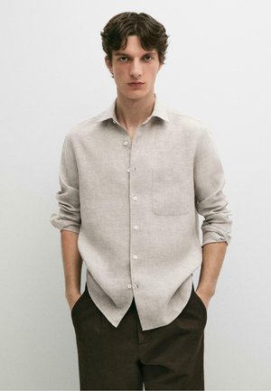 Junger Mann mit welligem dunklem Haar, trägt ein hellbeiges Hemd mit Knöpfen und hochgekrempelten Ärmeln sowie dunkelbraune Hose, steht mit den Händen in den Taschen.