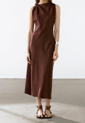 Burgundy, sleeveless dress with a fitted bodice and draped shoulder detail, falling to mid-calf; paired with brown strappy sandals.
