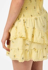 Vestido amarillo con volantes en capas y patrón floral. Presenta una tela texturizada y un borde de encaje a lo largo de los bordes. Toma cercana de la vista lateral.