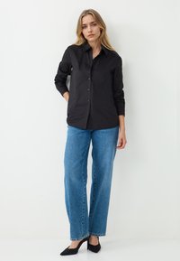 Zwarte blouse met lange mouwen, gecombineerd met blauwe wijde jeans en zwarte pumps met puntige neuzen. Glad materiaal en klassiek kraagontwerp.