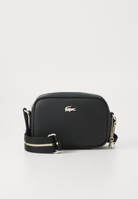 Crna kožna torba preko tijela s teksturiranom površinom, zatvaranjem na patentni zatvarač, srebrnim detaljima i crnim remenom s zlatnom prugom. Upečatljen Lacoste logo.