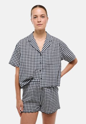Femme portant une chemise à manches courtes à carreaux noirs et blancs boutonnée et un short assorti aux bords festonnés, debout avec une main sur la hanche.