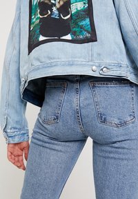Light blue denim jacket with a large graphic patch on the back, paired with fitted light blue jeans, showcasing a soft texture and classic cut.
