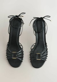 Paire de sandales plates en cuir noir avec plusieurs fines lanières, petites boucles à l'avant et liens autour de la cheville avec embouts métalliques, vues de dessus.