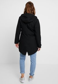 Giacca impermeabile nera con cappuccio, vita stretta e orlo posteriore più lungo. Indossata sopra jeans azzurri chiari e sneaker grigie.