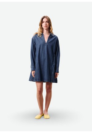 Jeune femme debout portant une robe bleue à fines rayures larges à manches longues et des chaussures plates jaunes sur un fond blanc uni.