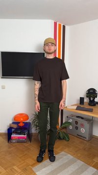 Brūns džemperis ar apaļu kaklu, olīvu zaļas bikses, melni sporta apavi. Istabā ir televizors, krāsaina siena un mēbeles neformālā stilā.