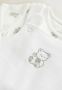 Weiße Baby-Latzhosen aus weichem Stoff mit einem Bärenstickdetail. Mit einem bedruckten Innenshirt mit kleinen Bärenmustern.