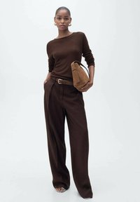 Un pull marron en tissu doux associé à un pantalon marron taille haute et large. Elle tient un petit sac en daim qui complète sa tenue.