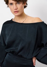 Zwarte, gestructureerde top met een off-shoulder ontwerp en een losse pasvorm. Bevat een brede halslijn en een geribbelde zoom, gecombineerd met een zilveren ketting.