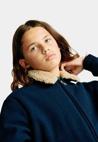 Granatowa kurtka polarowa z kołnierzem z kremowego sztucznego owczej wełny, z suwakiem i ściągaczami przy rękawach, zaprojektowana w luźnym kroju dla zapewnienia ciepła.