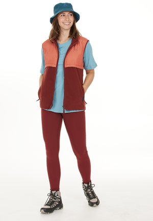 Fleeceväst i orange och vinröd, teal T-shirt, vinröda leggings och svarta vandringsskor med grå accenter, toppat med en blå bucket hat.