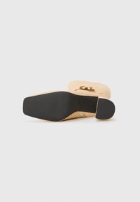 Chaussure à talon haut beige avec une semelle en caoutchouc noire, bout arrondi et accent en doré. Fini texturé sur la semelle pour une meilleure adhérence.