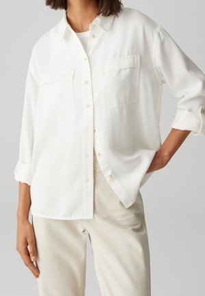 Witte blouse van zachte stof met twee borstzakken, opgerolde mouwen en een afgeronde zoom. Draagt bij lichtbeige broek.