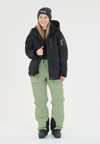 Zwarte geïsoleerde jas met een rits aan de voorkant en een logo patch op de mouw, gecombineerd met lichtgroene waterdichte broek en zwarte schoenen.