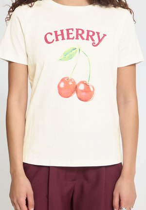 Personne portant un T-shirt crème avec le texte « "CHERRY" » et un motif de deux cerises rouges, assorti à un pantalon bordeaux.