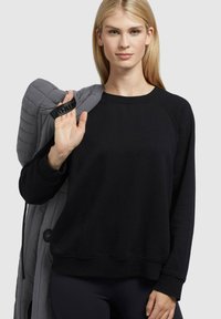 Blond kvinde iført sort sweater, som holder en grå quilted jakke over sin skulder med et håndtag, stående mod en ensfarvet lys baggrund.