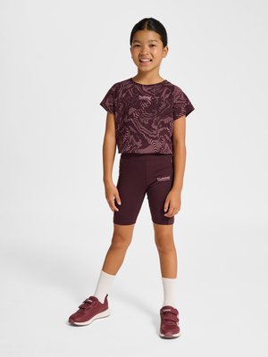 Młoda dziewczyna stoi i się uśmiecha, ubrana w bordowy strój sportowy z wzorzystym topem, szortami, białymi skarpetkami i pasującymi bordowymi trampkami.