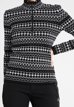 Czarno-biała bluzka w geometryczne wzory z długim rękawem, krótkim zamkiem i wysokim kołnierzem, zestawiona z czarnymi spodniami.