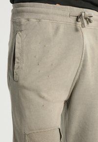 Beige Baumwoll-Jogginghosen mit einem Kordelzugbund, seitlichen Taschen und einem strukturierten Patch-Detail am Knie. Dezente Abnutzungsspuren auf dem Stoff.