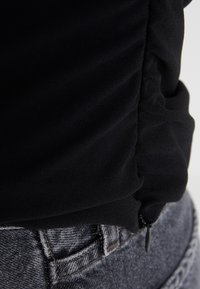 Tissu noir texturé avec côtés froncés, associé à un denim gris. Présente un petit détail de fermeture éclair sur le vêtement et des coutures visibles.