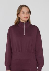 Pullover in tessuto viola scuro con colletto mezza zip, vestibilità morbida, maniche lunghe e vita elasticizzata per una finitura aderente.