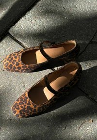 Bailarinas de charol con estampado de leopardo, con puntera cuadrada y correa en el tobillo, que presentan una textura suave y un interior en color marrón claro sobre un pavimento de piedra.