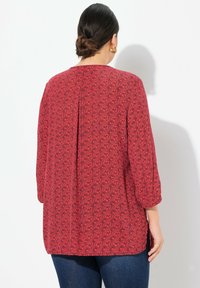 Rotes Tunika-Oberteil mit einem strukturierten Muster aus wirbelnden Designs, Dreiviertelärmeln und einer lockeren Passform mit seitlichen Schlitzen.