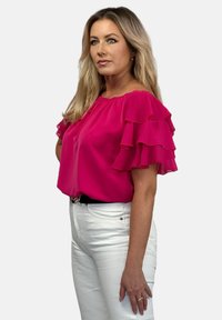 Różowa bluzka z warstwowymi falbanami na rękawach, wykonana z lekkiego materiału. Noszona z białymi dżinsami i czarnym paskiem, o dopasowanym kroju.