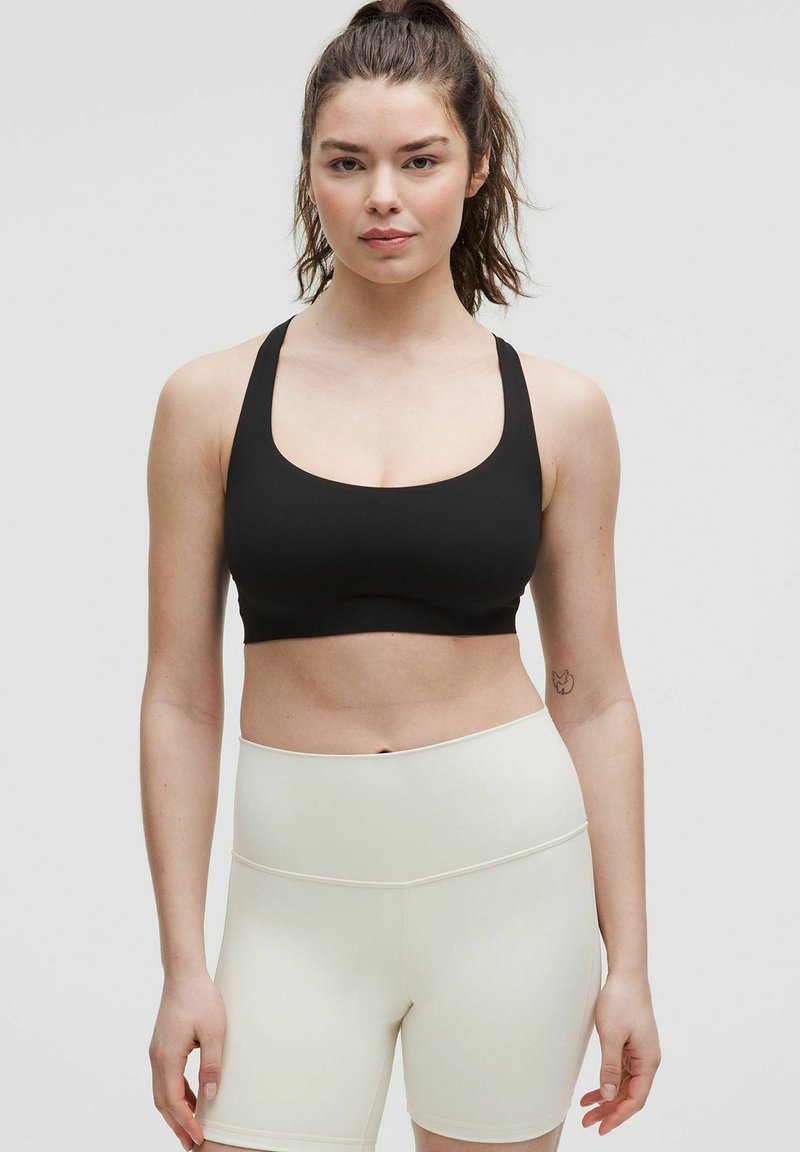 Sujeción deportiva negra con tirantes anchos, escote redondo y textura lisa, combinada con pantalones cortos de cintura alta de color claro.