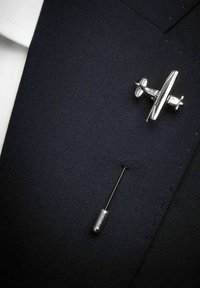 Silver flygplansnål och säkerhetsnål på en mörk texturerad tygbakgrund, som kontrasterar med en vit skjortkrage.