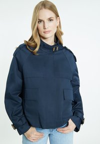 Navy sininen takki, jossa on korkea kaulus, suuret etutaskut ja muotoillut hartiat nappiaksentilla. Teksturoidusta kankaasta, lyhyt malli.