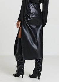 Jupe en cuir noire, élégante et taille haute, avec une fente latérale, associée à des bottes en cuir brillantes montantes à la cuisse, avec des pointes et un détail de zip.