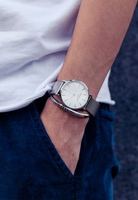 Reloj con correa de malla plateada y esfera redonda blanca, marcadores de hora negros y caja de metal pulido. Lleva sobre pantalones de pana azules.