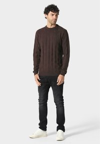 Brauner strukturierten Pullover mit geometrischem Muster, kombiniert mit dunkelblauen Jeans und weißen Sneakers. Einfacher runder Ausschnitt und lange Ärmel.