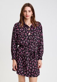Vestido camisero floral hecho de tejido ligero con un fondo negro y pequeñas flores rosas. Cuenta con un cuello tipo solapa y detalle de botones.