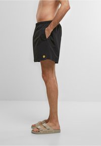 Sorte svøm shorts med elastisk talje, sidelommer og et gult logo. Båret med beige sandaler med et dobbelt spænde design.