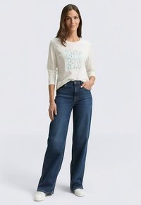 Long-sleeve cream top with teal abstract print, paired with high-waisted dark blue jeans and white sneakers, standing on a light background.