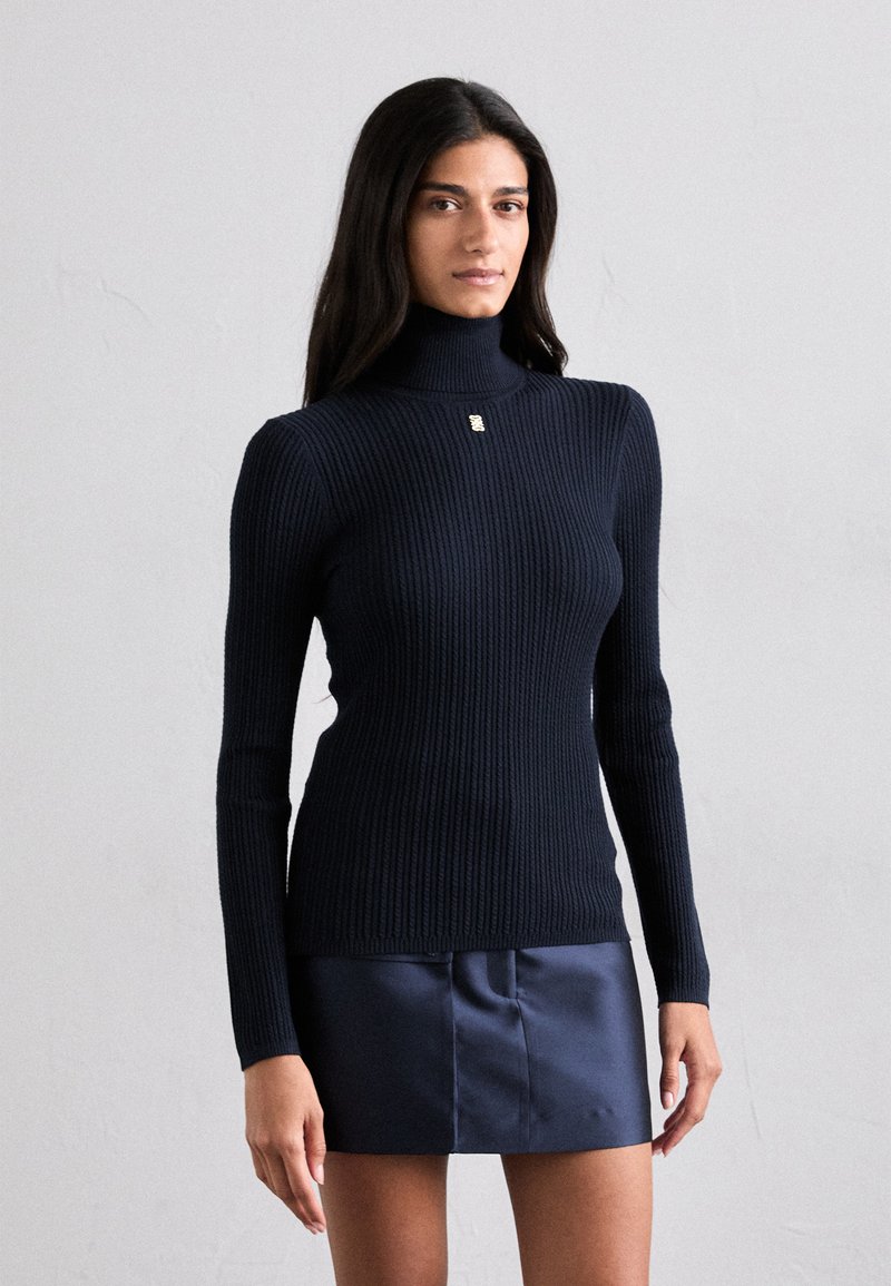 En kvinde iført en ribbet sort turtleneck sweater og en skinnende sort miniskirt foran en ensfarvet lys baggrund.