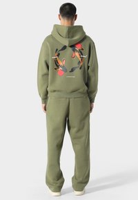 Oliivinvihreä huppari ja siihen sopivat housut. Takana on graafinen printti punaisilla ja mustilla elementeillä. Pehmeä pinta.
