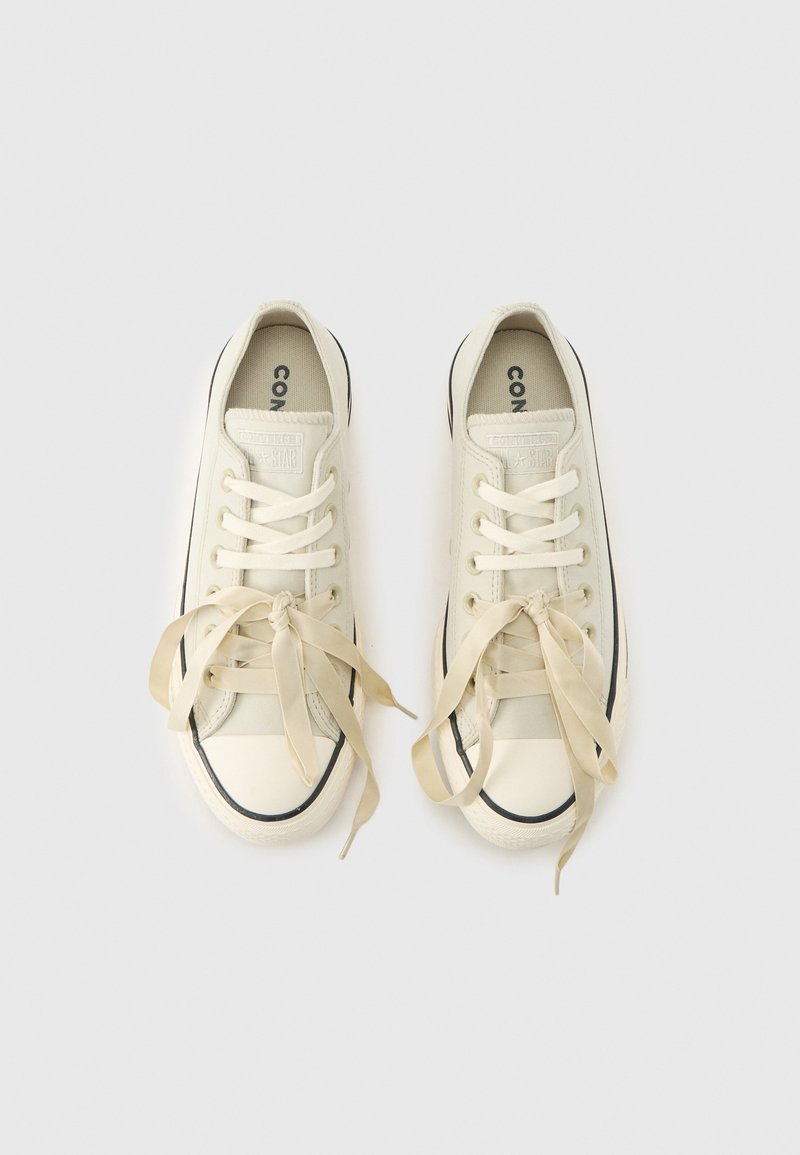 Sneakers basse in tela color avorio con lacci a nastro di raso e rifiniture nere, viste dall'alto su uno sfondo chiaro e uniforme.