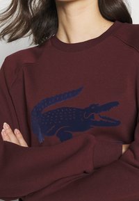 Sweatshirt bordeaux avec un grand motif crocodile bleu marine sur le devant. Col rond et manches raglan. Tissu doux au toucher.