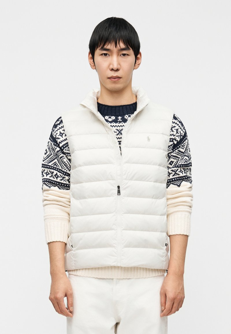 Ελαφρύ, λευκό γιλέκο με φερμουάρ και ψηλό γιακά π άνω από πουλόβερ με navy και κρεμ μοτίβο. Λείο ύφασμα, καπιτονέ σχέδιο.